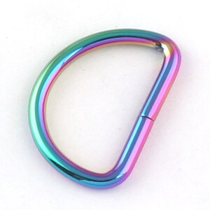 Puede incluir: Un anillo en D con un acabado iridiscente de arcoíris. Los herrajes metálicos tienen una superficie lisa y pulida con un espectro de colores que incluye azul, verde, morado y rosa. El anillo en D tiene forma de semicírculo con una barra recta.