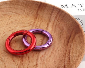 Può includere: Due portachiavi in metallo, uno rosso e uno viola, sono mostrati su una superficie di legno. Gli anelli sono aperti e pronti per essere utilizzati.