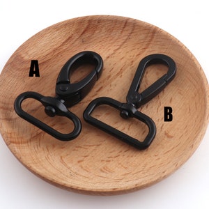 May include: Two black metal swivel clasps, labeled A and B, on a wooden surface. The clasps are used for attaching straps or chains to bags, purses, or other items.