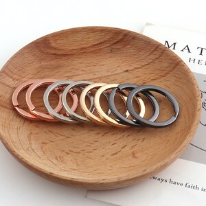 Puede incluir: Un juego de ocho llaveros en diferentes acabados metálicos, incluyendo plata, oro, oro rosa y negro. Los llaveros están dispuestos en círculo sobre una superficie de madera.