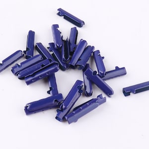 May include: A group of 18 blue metal cord ends, also known as aglets, for shoelaces or other cords. The cord ends are scattered on a white surface.