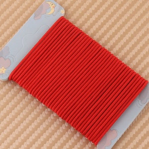 May include: A spool of bright red elastic cord, neatly wound and ready for use. The cord is tightly packed, showing a ribbed texture. The spool is attached to a light blue card with a cloud and moon design, suggesting a craft or sewing supply.