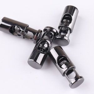 Puede incluir: Cinco topes de cordón de metal negro con un mecanismo de resorte. Los topes son cilíndricos y tienen un pequeño agujero en cada extremo.