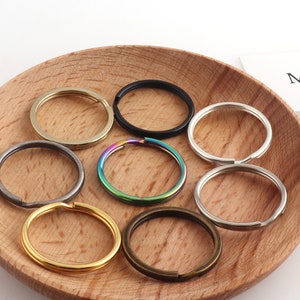 Könnte beinhalten: Sortiment von sieben Schlüsselringen in verschiedenen Farben und Ausführungen, darunter Gold, Silber, Schwarz und Regenbogen. Die Schlüsselringe werden auf einer Holzoberfläche präsentiert.