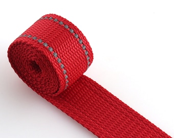 May include: Red webbing with a reflective gray stripe running along the edge. The webbing is rolled up and the end is laying flat.