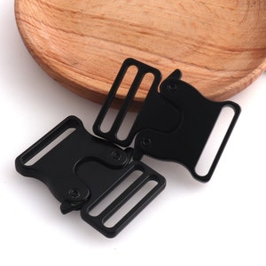 May include: Two black metal side release buckles. The buckles are connected together. The buckles are commonly used for straps, bags, and other accessories.