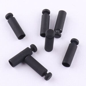 Puede incluir: Seis tapones de extremo de cable de plástico negro. Los tapones son cilíndricos con una parte superior redondeada y un agujero en el centro.