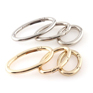 May include: A set of four oval-shaped metal clasps, two silver and two gold. The clasps are open and ready to be attached to a bag or other item.