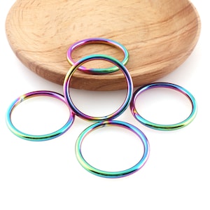 May include: Five iridescent metal rings, each with a different rainbow color pattern, are arranged on a white surface. The rings are all the same size and shape, and they are all made of a smooth, shiny metal.
