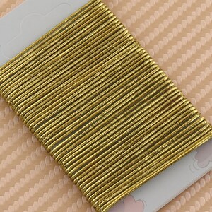 May include: A close-up of a gold elastic hair tie made of many thin strands of gold thread. The hair tie is on a white and pink background.