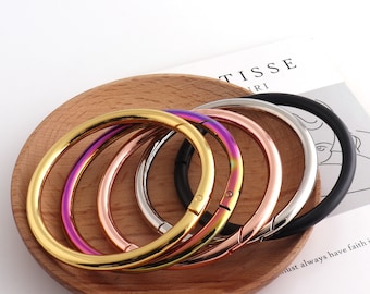 May include: Five different colored metal bangle bracelets with a hinged clasp. The bracelets are arranged in a circle on a wooden surface. The bracelets are gold, rose gold, silver, black, and iridescent.
