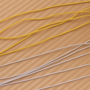 May include: A close-up of two types of elastic cord. One is yellow and the other is white. The cords are lying on a textured surface.