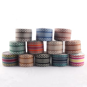 May include: A collection of 12 colourful striped woven ribbon trims. The trims are arranged in a semi-circle with the ends of each trim overlapping the next. The trims are all different colours and patterns, including brown, blue, red, green, yellow, and white.