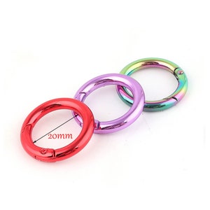 May include: Three round metal spring snap rings in red, purple, and iridescent rainbow colors. The red ring measures 20mm in diameter. These rings are designed to open and close with a spring mechanism.