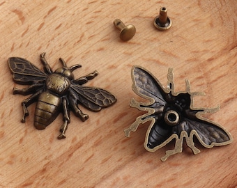 May include: Two antique gold-toned bee charms with a detailed design. One bee charm is solid and the other is an outline. Two small antique gold-toned rivets are also included.