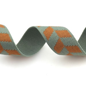 May include: A green and orange patterned woven ribbon. The ribbon is woven with a repeating pattern of orange squares on a green background.
