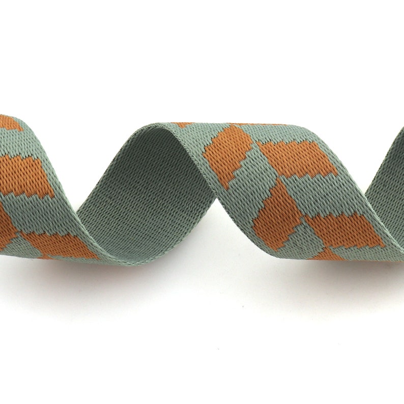 May include: A green and orange patterned woven ribbon. The ribbon is woven with a repeating pattern of orange squares on a green background.