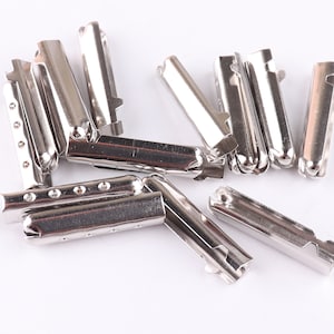 May include: A group of silver metal garment end caps, also known as  'cord ends' or 'cord tips', are scattered on a white surface. These metal caps are used to finish the ends of cords or straps.