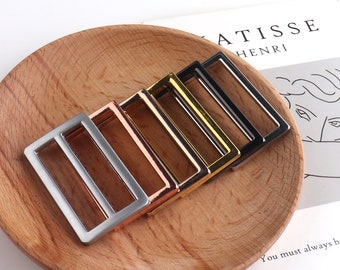 May include: Five metal slider buckles in different finishes: silver, rose gold, gold, brass, and black. The buckles are arranged in a row on a wooden surface.
