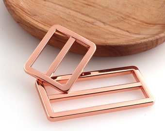 May include: Two rose gold metal slider buckles. The larger buckle is rectangular with two horizontal bars. The smaller buckle is square with one horizontal bar.