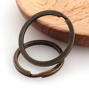 May include: Two antique gold-toned metal key rings. The larger ring is slightly open and the smaller ring is closed. The rings are on a wooden surface.