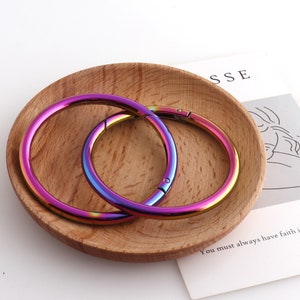 May include: Two iridescent rainbow colored metal rings with a spring closure. The rings are on a wooden surface.