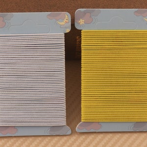 May include: Two packs of hair elastics, one white and one yellow, are displayed on a brown background. Each pack has a cartoon design of a moon and stars.