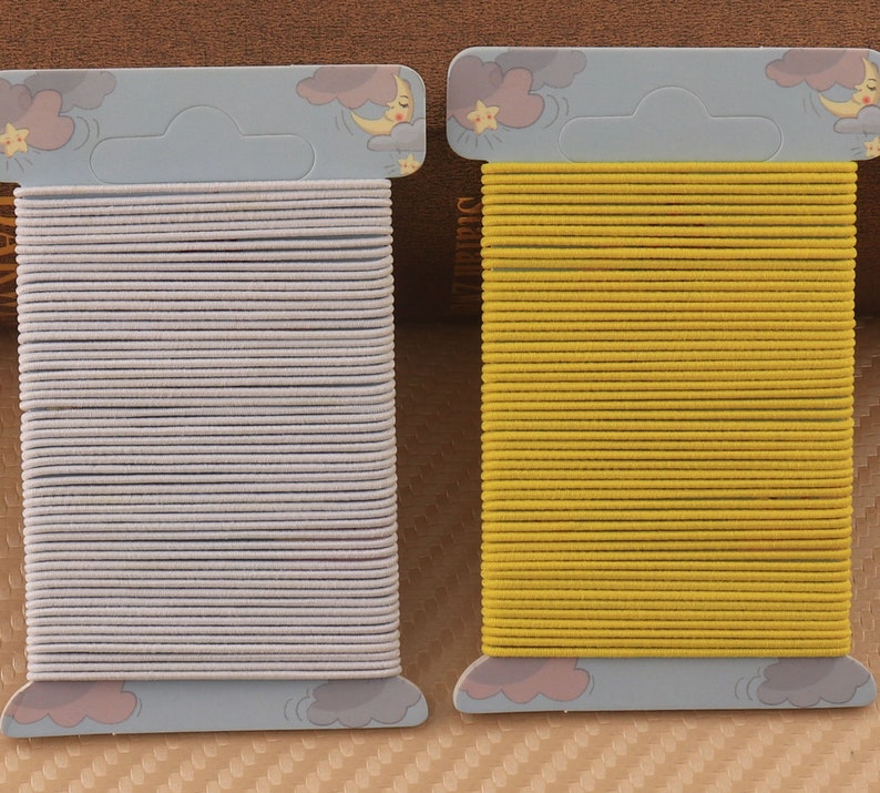 May include: Two packs of hair elastics, one white and one yellow, are displayed on a brown background. Each pack has a cartoon design of a moon and stars.