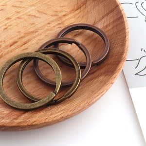 May include: Close-up of three metal key rings in antique brass and bronze tones. The key rings are open, ready to attach keys. The rings are on a wooden plate.