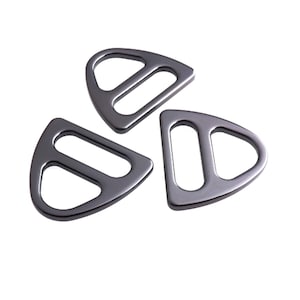 Pode incluir: Três fivelas metálicas cinza-chumbo, com formato triangular e duas aberturas. Apresentadas sobre um fundo branco, destacando o seu design e acabamento. Estas fivelas são provavelmente usadas em bolsas ou outros acessórios.
