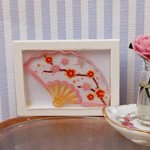 May include: A miniature white framed artwork featuring a pink and yellow fan design with red and pink floral accents. A small glass vase with pink roses and a floral patterned dish are also present.