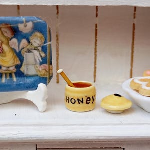 以下が含まれることがあります： 天使が描かれた青いトレイ、木製のディッパーが付いた小さな黄色の蜂蜜ポット、ミニチュアパイが特徴のミニチュアシーン。 蜂蜜ポットには黒字で「HONEY」と書かれています。 パイは黄色で黒い点があります。