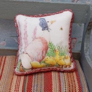May include: A miniature decorative pillow with a charming illustration of a goose, ducklings, and a butterfly. The pillow has a white background with a pink border and is placed on a striped fabric.