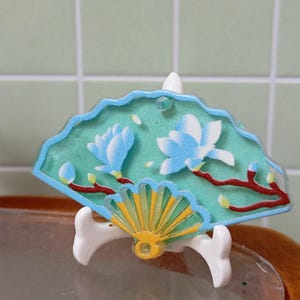May include: A decorative fan with a teal background and a light blue border. The fan features a floral design with light blue flowers and red branches. The fan is supported by a white stand with a gold fan base.