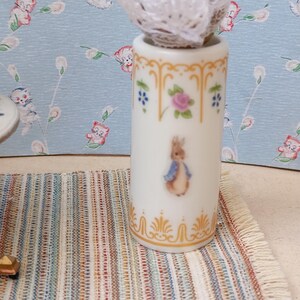 Op de afbeelding: Een witte keramische vaas met een Peter Konijn-illustratie, versierd met gouden accenten, roze rozen en blauwe bloemmotieven. De cilindrische vaas staat op een gestreept tapijt. Een kleine tafel met een bloemmotief is zichtbaar.