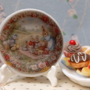 May include: A small, decorative ceramic plate featuring a picnic scene with rabbits. The plate has a floral border and is displayed on a white stand. A small plate of miniature cakes and donuts is next to it.