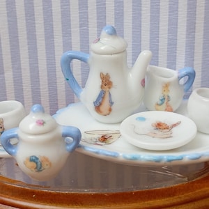 Può includere: Un servizio da tè in miniatura in porcellana con teiera, lattiera, zuccheriera, due tazze e due piattini. Il set è bianco con dettagli blu e illustrazioni di un coniglio. Il set è presentato su un vassoio bianco con bordatura blu.