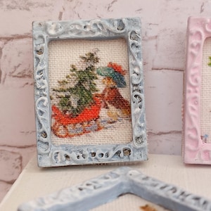 Op de afbeelding: Miniatuur ingelijste kruissteekkunst met een wintertafereel met een kerstboom en een persoon in een slee. De lijst is lichtblauw met sierlijke details. Het kunstwerk staat op een crèmekleurige achtergrond.