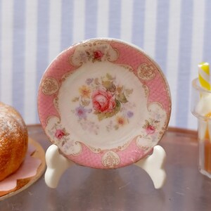 May include: A miniature pink and white floral plate with a rose design, displayed on a white stand. The plate is part of a miniature tea set, with a cake and a sundae in the background.