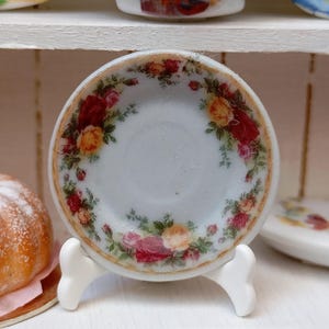 Pode incluir: Um pequeno prato de porcelana branca com um desenho floral de rosas vermelhas, amarelas e rosas. O prato tem uma borda dourada e é exibido em um suporte branco. Parte de uma coleção em miniatura.