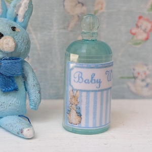 May include: A light blue miniature rabbit figurine with a matching blue bow tie next to a small, translucent teal bottle. The bottle features a white label with blue vertical stripes, the text Baby W, and a small illustration of a rabbit.