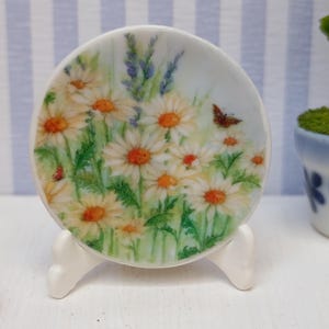 Könnte beinhalten: Ein kleiner, weißer Keramikteller mit einem Blumendesign, das Gänseblümchen, grüne Blätter und einen Schmetterling zeigt. Der Teller wird von einem weißen Ständer getragen. Der Hintergrund umfasst ein blau-weiß gestreiftes Muster und eine kleine Topfpflanze.