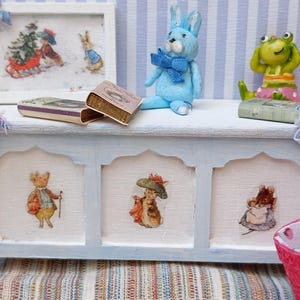 May include: A miniature, pale blue chest with three panels, each featuring a different animal illustration. A blue stuffed bunny, a green frog figurine, and two small books sit on top. A framed picture and a pink basket are also visible.