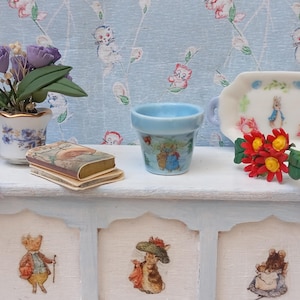 May include: A miniature scene featuring a small blue flower pot with a Peter Rabbit design, a tiny book, a white tray with a Peter Rabbit illustration, and a pot of purple flowers. The scene is set on a light blue painted chest.