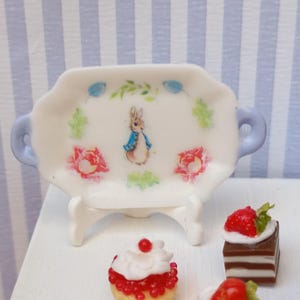 Può includere: Un vassoio bianco in miniatura con bordo smerlato e manici azzurri. Il vassoio presenta un'illustrazione di Peter Rabbit e decorazioni floreali. I dessert in miniatura, tra cui torte, sono in primo piano.