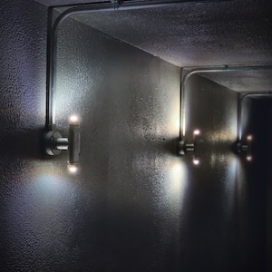 Puede incluir: Un pasillo oscuro con una serie de apliques de pared plateados con dos luces cada uno. Las luces están encendidas e iluminan el pasillo.