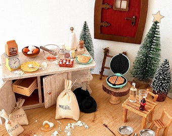 May include: A miniature kitchen scene featuring a red arched door, a wooden counter with baking supplies, a waffle maker on a pedestal, small evergreen trees, a wooden table with chairs, a scale labeled EPICERIE, and gingerbread figures.