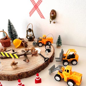 Pu&ograve; includere: Una scena di cantiere in miniatura con giocattoli gialli, coni stradali e una pala, disposti su una fetta di legno. La scena comprende alberi in miniatura e un casco da cantiere, creando un tema di costruzione giocoso.