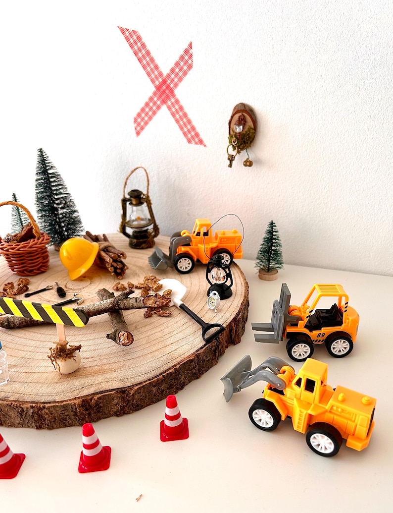 Pu&ograve; includere: Una scena di cantiere in miniatura con giocattoli gialli, coni stradali e una pala, disposti su una fetta di legno. La scena comprende alberi in miniatura e un casco da cantiere, creando un tema di costruzione giocoso.