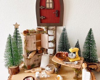 Può includere: Un set da cucina per fate in miniatura con una porta rossa, un armadio in legno, un'impastatrice gialla, un tavolo in legno e un sacco di farina. Il set è circondato da alberi di pino in miniatura e una base in legno.